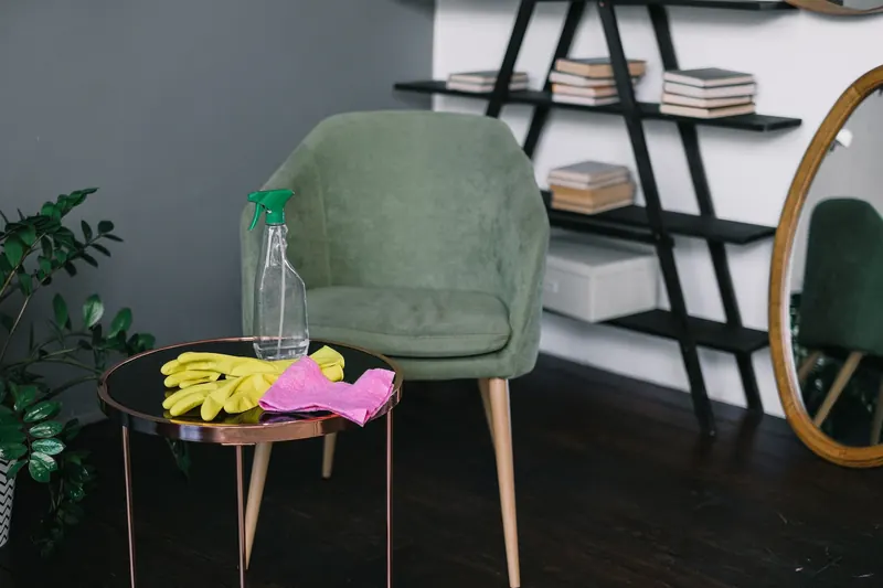 Yellow latex gloves placed near spray detergent bottle and rag on round table against comfortable chair in living room in daytime
