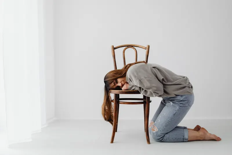 A woman kneels and rests on a wooden chair in a minimalist setting, conveying tranquility