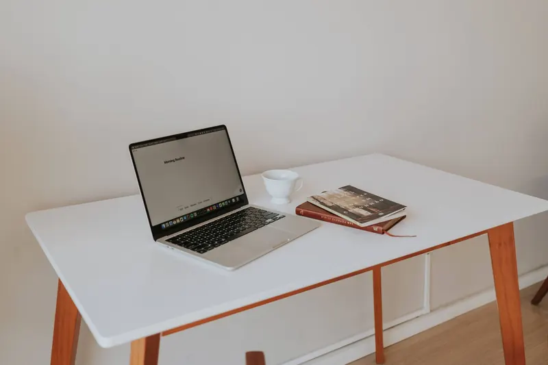 A minimalistic workspace featuring a laptop, coffee cup, and books on a white desk with wooden legs
