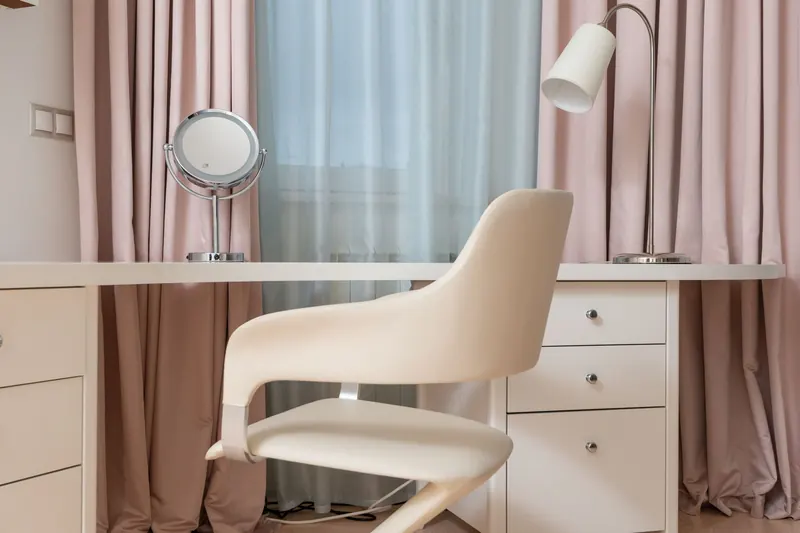Contemporary white chair near table with cabinet and lamp near window with pink curtains