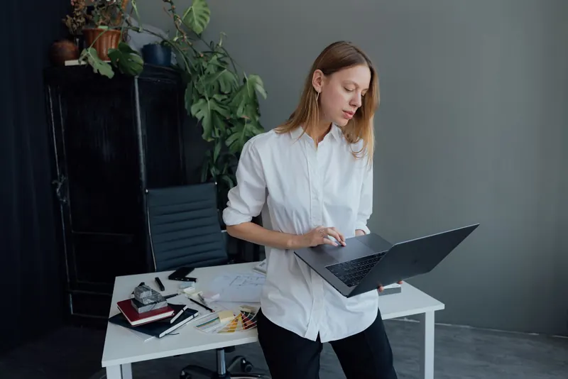 Young woman in white shirt working from home on a laptop, standing at a desk with plants